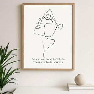 Può includere: Una stampa artistica incorniciata con un disegno a linea minimalista nero di un volto su sfondo bianco. L'opera d'arte è in una cornice di legno marrone chiaro. La stampa include il testo: "Be who you came here to be. The rest unfolds naturally."