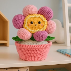 Könnte beinhalten: Eine handgefertigte Häkelblume in einem rosa Topf. Die Blume hat eine gelbe Mitte mit einem lächelnden Gesicht, umgeben von rosa und lila Blütenblättern. Grüne Blätter und eine graue Basis vervollständigen das Design. Ein charmantes, skurriles Dekorationsstück.