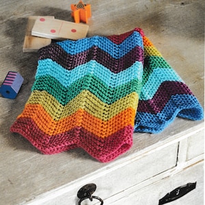 Puede incluir: Una manta tejida con un patrón de chevrón en colores del arcoíris, incluyendo rojo, naranja, amarillo, verde, azul y morado. La manta está doblada sobre una superficie de madera, con juguetes de madera en el fondo.