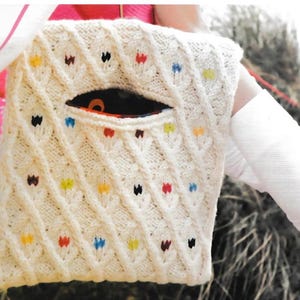 May include: Cream-colored knitted bag with a diamond pattern and colorful accents. The bag has an oval opening and is held by a person wearing a white long-sleeved shirt.