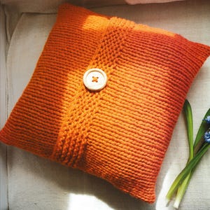 以下が含まれることがあります： 大きな木製のボタン留めが付いた、オレンジ色のニットの正方形のクッション。 クッションは、質感のあるリブ編みのデザインです。 明るい色の表面に置かれています。 小さな青い花が付いた緑色の茎がいくつか前景にあります。