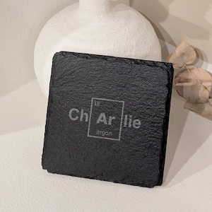 Könnte beinhalten: Schwarzer Schieferuntersetzer mit dem Text "Charlie" und dem chemischen Symbol für Argon, "Ar", mit der Ordnungszahl 18.