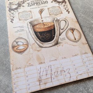 以下が含まれることがあります： コーヒーをテーマにしたカレンダー。エスプレッソショットのイラストとラベル、テキストが特徴です。カレンダーはスパイラル綴じで、吊り下げ用の穴があります。3月が表示されています。