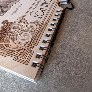 以下が含まれることがあります： モカポットとコーヒー豆の詳細なイラストが描かれた、スパイラル綴じのノート。表紙には「Der Moka Pot」と「Der italienische Klassiker f&uuml;r den Herd」の文字がヴィンテージスタイルで表示されています。
