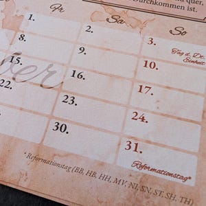 Op de afbeelding: Een beige kalenderpagina met een vintage perkamenttextuur met een raster van genummerde dagen. De tekst bevat Duitse koppen zoals Fr, Sa en So, samen met een kader met Duitse tekst over verdediging en de feestdag Reformationstag.