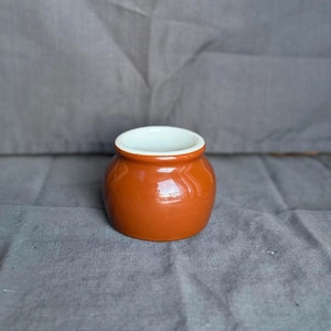 Può includere: Un piccolo vaso rotondo in ceramica con esterno arancione-marrone lucido e interno bianco. Il vaso ha un'ampia apertura e un design semplice. Probabilmente viene utilizzato per la conservazione o la decorazione.