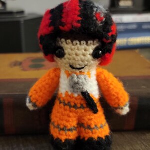 May include: A small, handmade crochet doll featuring a pilot wearing a bright orange flight suit with grey accents, black boots, and a red and black helmet. The figure has black bead eyes, a stitched smile, and a small grey microphone accessory.