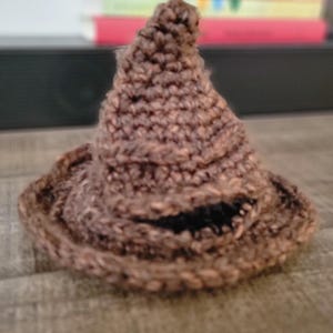 May include: A small, brown, crocheted hat with a pointed, slightly slumped top and a wide, circular brim. The yarn texture is visible throughout the piece, featuring a dark, horizontal opening near the base that resembles a mouth-like fold.