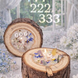 Könnte beinhalten: Zwei rustikale Kerzenhalter aus Holz mit brennenden Kerzen, verziert mit Kristallen, Blumen und den Zahlen 777, 222 und 333. Die Kerzen stehen auf weißem Sand und schaffen eine ruhige und spirituelle Atmosphäre.
