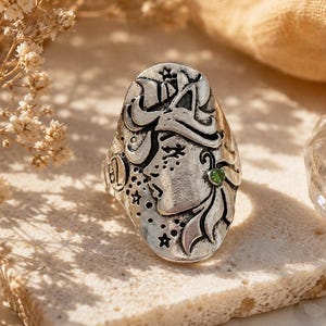 Puede incluir: Un anillo de tono plateado con una cara ovalada que presenta el perfil en relieve de una persona, motivos de estrellas y el símbolo del zodiaco Leo. Una pequeña piedra preciosa redonda de color verde está engastada en el diseño lateral.