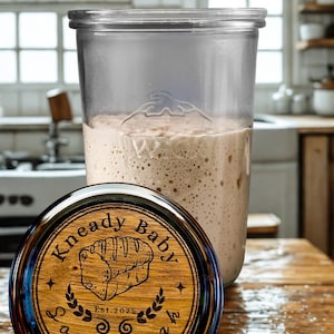 May include: A clear glass jar with a light-colored, bubbly substance inside, likely sourdough starter. The jar is on a wooden surface, with a wooden lid that reads "Kneady Baby Sourdough" and features a bread illustration. The background shows a kitchen.