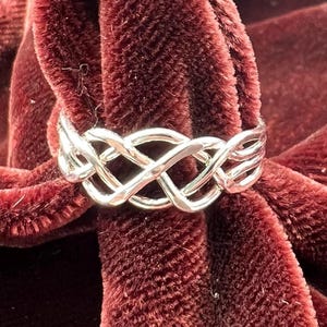 May include: A silver-colored ring with an intricate Celtic knot design. The ring is displayed against a backdrop of deep red velvet fabric, creating a striking contrast. The interwoven pattern is a prominent feature.