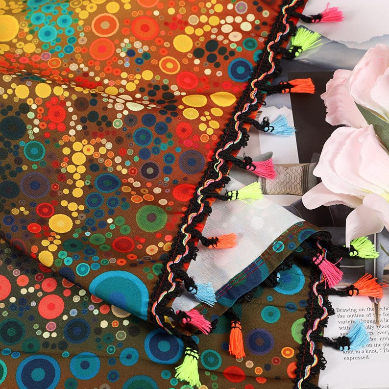 May include: A colorful scarf with a brown background and a pattern of circles in various colors. The scarf is trimmed with black fringe and colorful tassels in shades of pink, orange, blue, and green. The scarf has a decorative edge.
