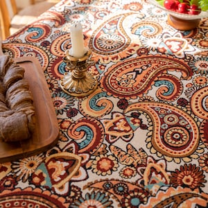 May include: A close-up of a paisley patterned tablecloth in shades of orange, brown, and teal. A gold candle holder with a white candle sits on the tablecloth. A wooden serving board with bread is on the left.