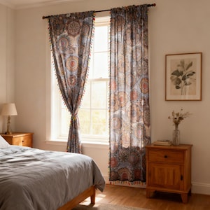 May include: A pair of patterned curtains with a blue background and circular designs in shades of orange, red, and brown. The curtains are trimmed with colorful tassels and hang in front of a window in a bedroom setting.