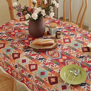May include: A colorful tablecloth with a geometric pattern in red, orange, and blue covers a wooden table. A vase of flowers, a loaf of bread, and a green plate with silverware are on the table. The tablecloth has a fringed edge.