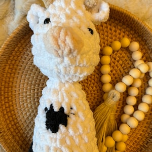 May include: White crocheted cow plush toy with black spots, sitting in a wicker basket with a wooden bead garland and a tan tassel.