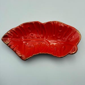 May include: A glossy, bright red ceramic dish shaped like a monstera leaf. The dish features a textured surface with embossed veins and perforations, finished with metallic gold paint along the scalloped edges and raised details for contrast.