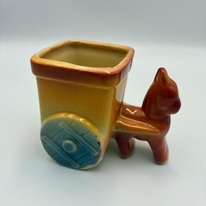 May include: A ceramic planter shaped like a donkey pulling a cart. The donkey is reddish-brown, while the cart body is yellow with a reddish-brown rim. The cart features a blue wheel with a textured spoke design and a hollow, open-top container.