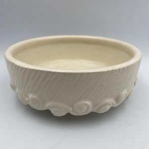 May include: Off-white ceramic bowl with a textured, diagonal pattern on the sides. The base features a decorative, repeating swirl design. The bowl has a smooth, rounded interior.