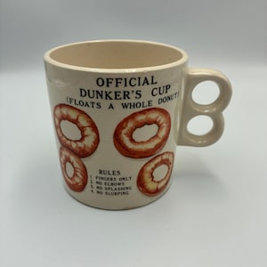 May include: A cream-colored ceramic mug with a unique double-loop handle. The front features the text OFFICIAL DUNKER'S CUP (FLOATS A WHOLE DONUT) above four illustrations of donuts and a list of rules: 1. FINGERS ONLY, 2. NO ELBOWS, 3. NO SPLASHING, 4. NO SLURPING.