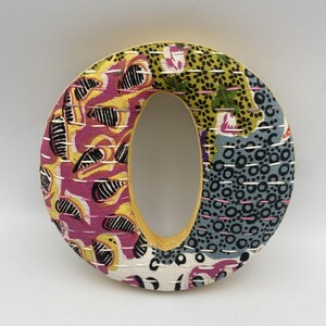 May include: A decorative letter "O" with a colorful fabric design. The letter features a yellow border and is covered in patterned fabric with pink, yellow, black, and blue hues. The design includes floral and abstract shapes.