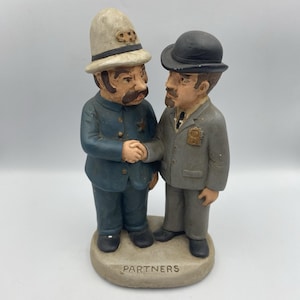 May include: A ceramic figurine depicting two men shaking hands. One figure wears a blue uniform and a helmet, the other a gray suit and bowler hat. The base reads "PARTNERS".