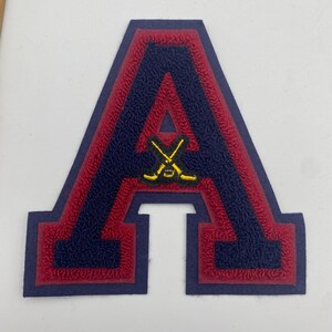 May include: A varsity-style letter A patch featuring a textured navy blue center with a red border and a dark blue outer edge. Centered on the letter is a small, embroidered yellow graphic of two crossed hockey sticks and a hockey puck.