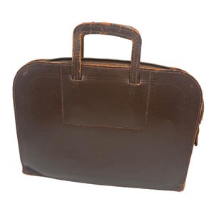 May include: A dark brown briefcase with a rectangular body and rounded top corners. It features a top zipper closure and two integrated handles showing signs of surface wear and scuffing on the edges. The front panel has a stitched rectangular detail.