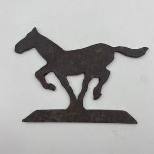 May include: A dark brown, weathered metal silhouette of a horse in a galloping pose, mounted on a flat, rectangular base. The surface of the metal shows signs of oxidation and a rough, textured finish throughout the entire horse figure.