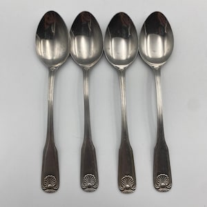 May include: A set of four silver-toned metal spoons arranged vertically. Each spoon features a polished oval bowl and a long, slender handle with a vertical ribbed pattern and a decorative shell-like motif at the base of the handle.