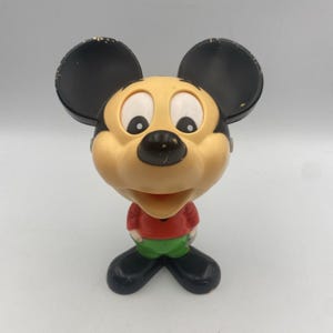 May include: A plastic Mickey Mouse figurine featuring a tan face, large black ears with visible paint wear, a red shirt, green pants, and black shoes. The character has white eyes with black pupils and a black nose, standing in a neutral pose.