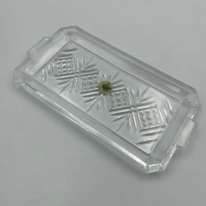 May include: A clear, rectangular glass tray with etched geometric designs and handles. The tray features a central diamond pattern with radiating lines. A small, round, green object rests in the center of the tray.