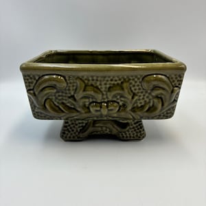 May include: A rectangular olive green ceramic planter featuring a raised scrollwork relief pattern on the sides and a textured stippled background. The vessel is supported by a central pedestal base and has a smooth, glossy glazed finish.