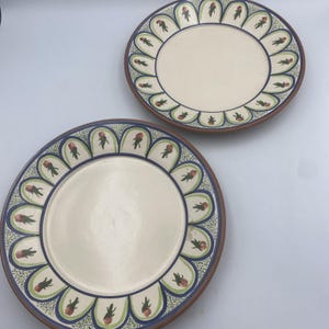 May include: Two ceramic plates with a cream-colored center and a decorative border. The border features a blue and green design with red floral accents. The plates have a brown rim.