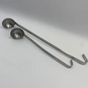 May include: Two stainless steel ladles with long handles and curved hooks. The round bowls are positioned at the top of the image, with the handles extending diagonally. The ladles are designed for cooking and serving soups, sauces, or other liquids.