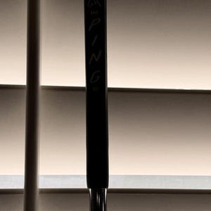 May include: A black golf club with the brand name "PING" vertically printed in white. The club features a black grip and a silver shaft. The top of the club has a logo of a bird.