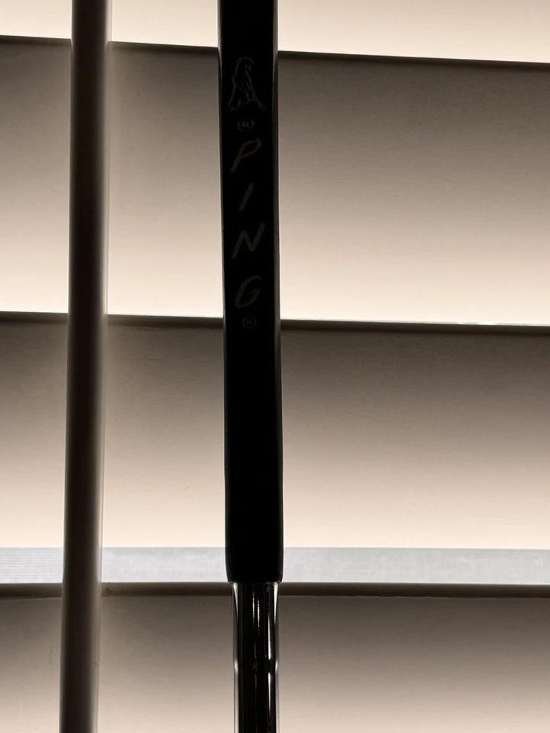 May include: A black golf club with the brand name "PING" vertically printed in white. The club features a black grip and a silver shaft. The top of the club has a logo of a bird.