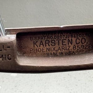 May include: Close-up of a vintage golf putter head. The putter is bronze-colored with the text "BALL-NAMIC" on the side. The center of the putter head has the text "KARSTEN CO. PHOENIX, ARIZ. 85029 MADE IN U.S.A."