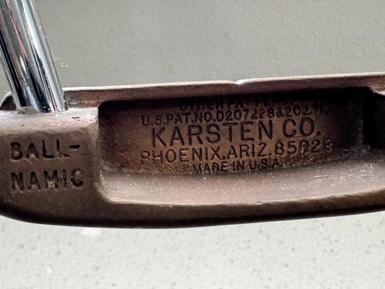 May include: Close-up of a vintage golf putter head. The putter is bronze-colored with the text "BALL-NAMIC" on the side. The center of the putter head has the text "KARSTEN CO. PHOENIX, ARIZ. 85029 MADE IN U.S.A."