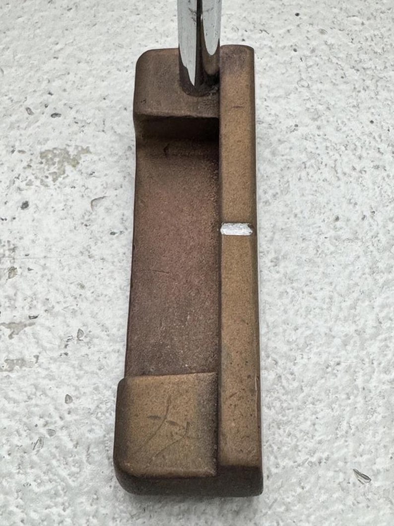 May include: A bronze-colored golf putter with a silver shaft. The putter head has a rectangular shape with a small white line. The putter has a worn, textured appearance.