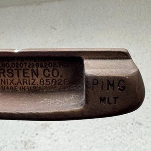 May include: Close-up of a vintage golf club head. The club is a rusty brown color and features the text "KARSTEN CO. PHOENIX, ARIZ. 85029 MADE IN U.S.A." and "PING MLT".