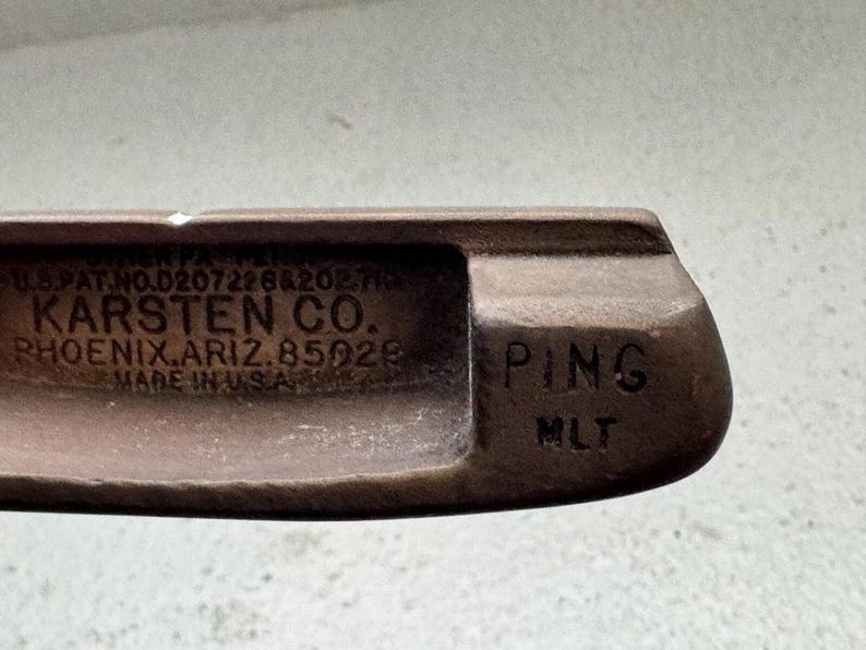 May include: Close-up of a vintage golf club head. The club is a rusty brown color and features the text "KARSTEN CO. PHOENIX, ARIZ. 85029 MADE IN U.S.A." and "PING MLT".