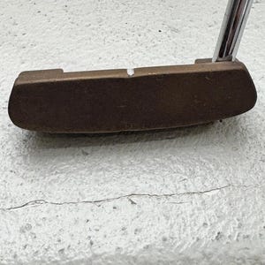 May include: A bronze-colored golf putter with a silver shaft. The putter head is rectangular with rounded edges and a small white mark. The shaft is angled upwards, suggesting a ready-to-use position. The putter is designed for precision and control on the green.