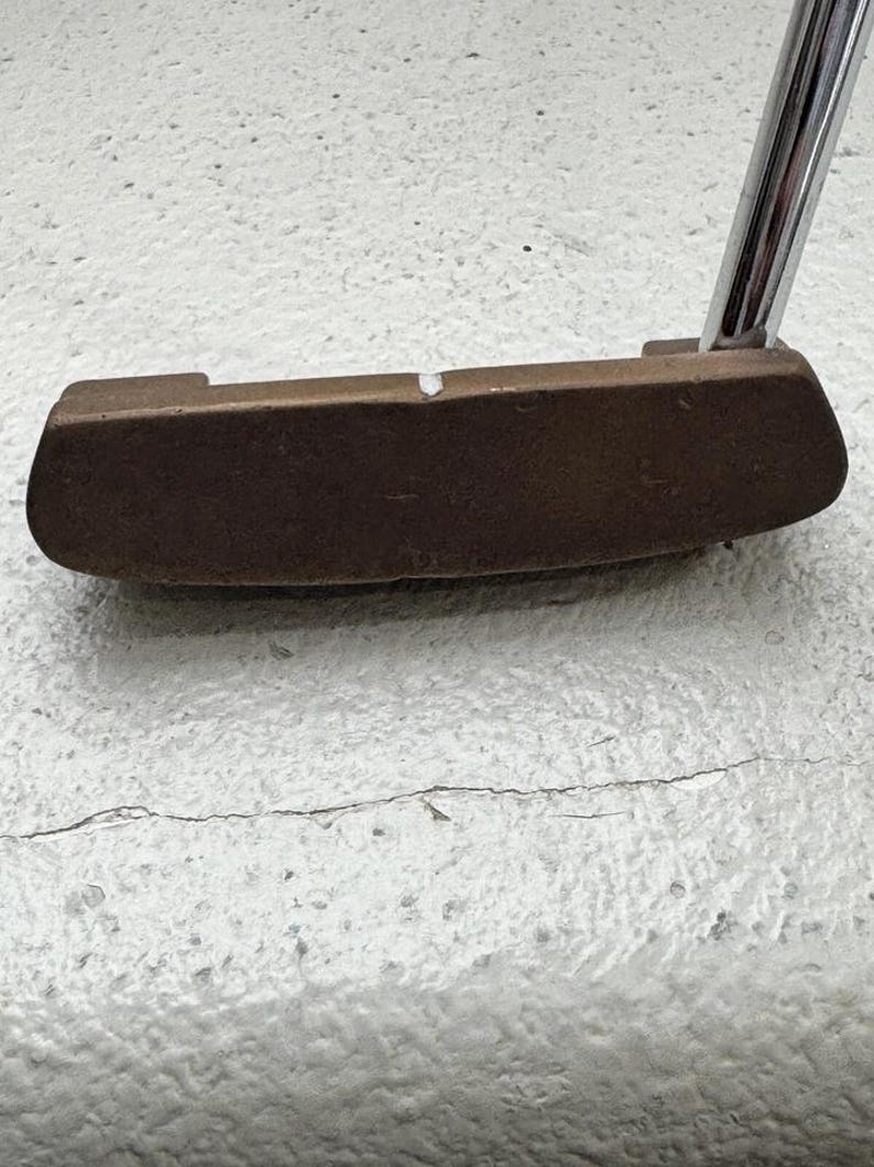 May include: A bronze-colored golf putter with a silver shaft. The putter head is rectangular with rounded edges and a small white mark. The shaft is angled upwards, suggesting a ready-to-use position. The putter is designed for precision and control on the green.