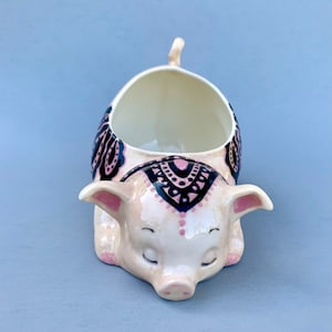 May include: A ceramic pig planter with a pink and black mandala design. The pig is lying down with its eyes closed and has a small opening on its back for planting.