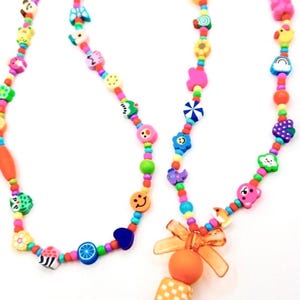 May include: A lanyard featuring a long string of multicolored beads in various shapes, including fruits, flowers, animals, and smiley faces. The bottom section includes an orange translucent bow, a round orange bead, and a square bead, ending in a metal clasp.