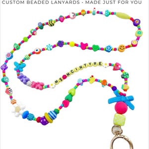 May include: A custom beaded lanyard featuring the text MS MCINTYRE, adorned with a variety of colorful shapes including flowers, butterflies, hearts, smiley faces, and gummy bears, finished with a gold-toned metal lobster clasp and a blue bow.
