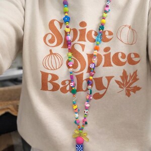 May include: A beige sweatshirt featuring the orange text Spice Spice Baby with pumpkin and leaf graphics, paired with a long, colorful beaded lanyard. The lanyard consists of various multi-colored plastic beads in shapes like flowers, hearts, and fruits, ending in a yellow bow and a metal clip.