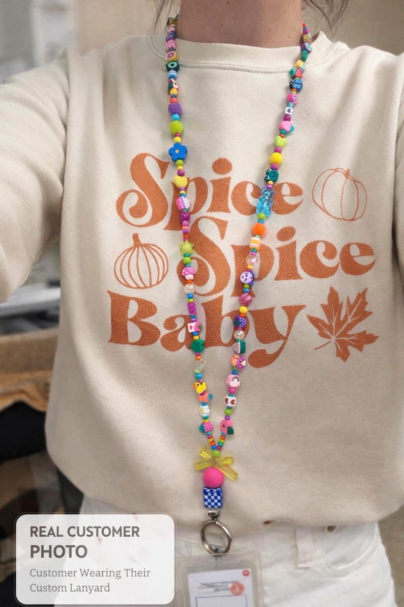 May include: A beige sweatshirt featuring the orange text Spice Spice Baby with pumpkin and leaf graphics, paired with a long, colorful beaded lanyard. The lanyard consists of various multi-colored plastic beads in shapes like flowers, hearts, and fruits, ending in a yellow bow and a metal clip.